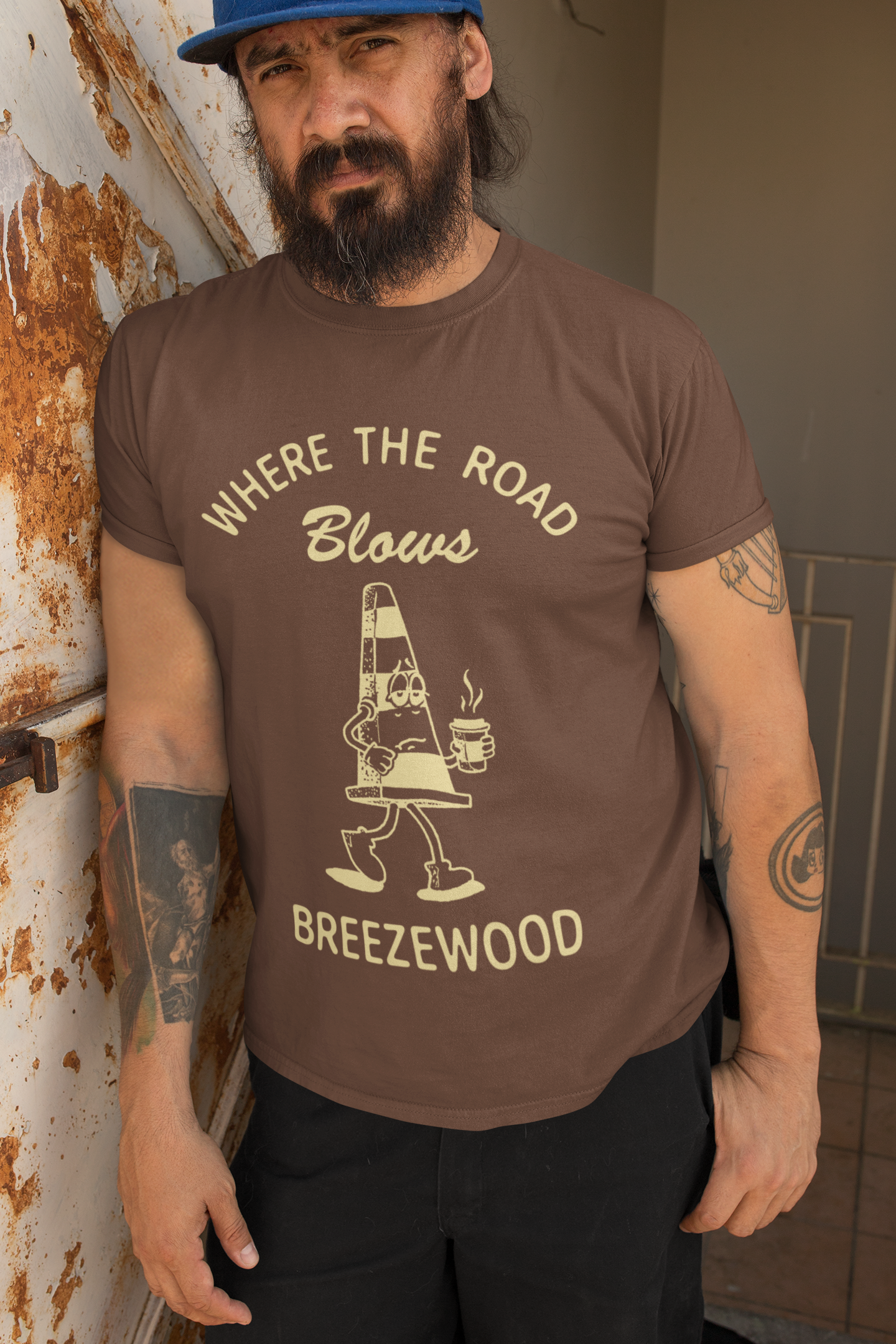 A bearded man wearing a brown t-shirt with a tired looking illustration of a traffic cone holding a cup of coffee that reads "Where the road blows - Breezewood" 