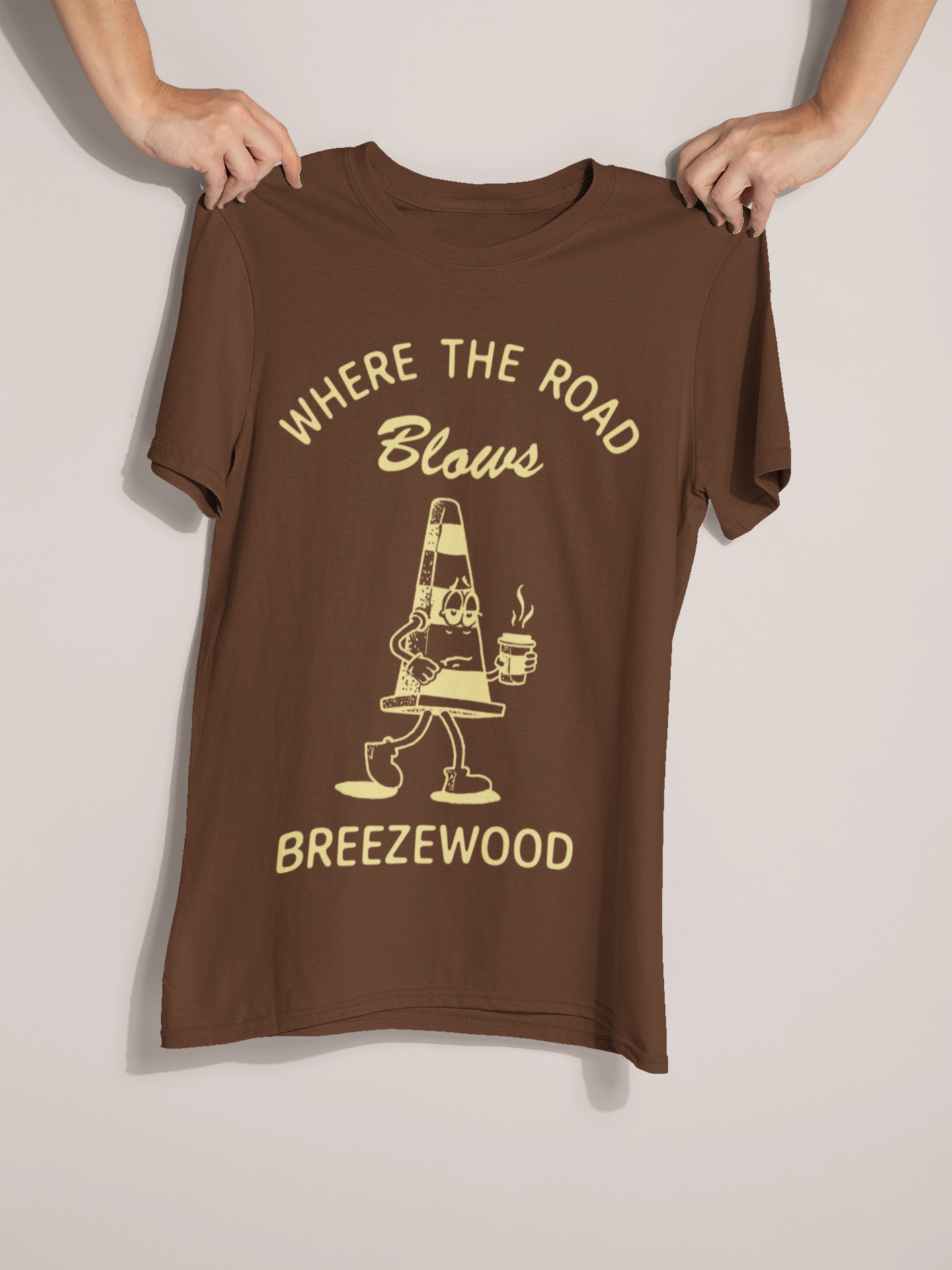 Hands holding brown t-shirt with a tired looking illustration of a traffic cone holding a cup of coffee that reads "Where the road blows - Breezewood" 